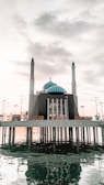 The image depicts a mosque named 'Masjid Amirul Mukminin' which is situated on stilts over a body of water. The mosque includes a central large blue-domed structure flanked by two tall minarets. Surrounding the mosque, several lampposts are visible, and the scene is set under a partly cloudy sky, creating reflections in the water below.