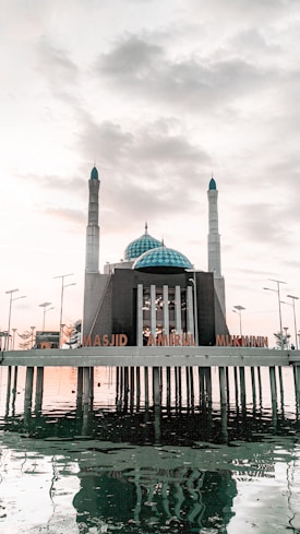 The image depicts a mosque named 'Masjid Amirul Mukminin' which is situated on stilts over a body of water. The mosque includes a central large blue-domed structure flanked by two tall minarets. Surrounding the mosque, several lampposts are visible, and the scene is set under a partly cloudy sky, creating reflections in the water below.