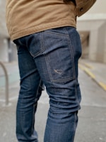 A person is wearing dark blue denim jeans and a tan jacket while standing on a pavement. The focus is on the lower half of the person, highlighting the textured fabric and stitching of the jeans. The background appears to be an urban setting with a blurred building and a railing.