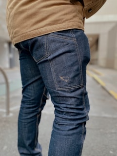 A person is wearing dark blue denim jeans and a tan jacket while standing on a pavement. The focus is on the lower half of the person, highlighting the textured fabric and stitching of the jeans. The background appears to be an urban setting with a blurred building and a railing.