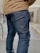 A person is wearing dark blue denim jeans and a tan jacket while standing on a pavement. The focus is on the lower half of the person, highlighting the textured fabric and stitching of the jeans. The background appears to be an urban setting with a blurred building and a railing.