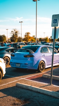 A light blue car is parked in an outdoor parking lot, surrounded by other vehicles. The scene is set during daylight with clear skies and scattered clouds. A reserved parking sign is seen in the foreground, and trees and additional streetlights can be observed in the background.