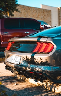 A sleek silver car is parked in a sunlit area, casting clear shadows onto the ground. The vehicle features distinctive rear tail lights and a visible license plate from Texas. Nearby, a red truck is partially visible, suggesting an urban or suburban parking lot setting.