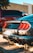 A sleek silver car is parked in a sunlit area, casting clear shadows onto the ground. The vehicle features distinctive rear tail lights and a visible license plate from Texas. Nearby, a red truck is partially visible, suggesting an urban or suburban parking lot setting.