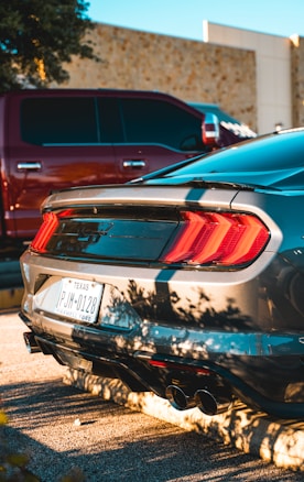 A sleek silver car is parked in a sunlit area, casting clear shadows onto the ground. The vehicle features distinctive rear tail lights and a visible license plate from Texas. Nearby, a red truck is partially visible, suggesting an urban or suburban parking lot setting.