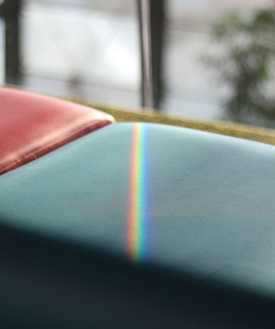 A soft, colorful gradient of a rainbow is visible on a smooth, light blue surface. In the background, there is an out-of-focus area with a hint of greenery and diffuse light entering through a window or reflective surface.