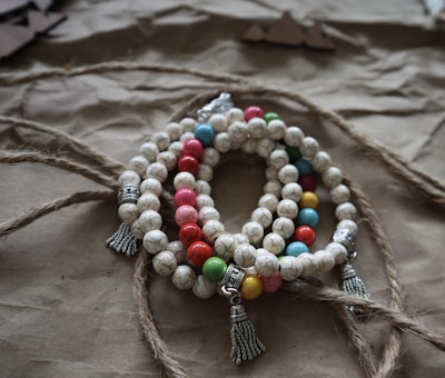 A set of beaded bracelets with multicolored spherical beads interspersed with larger, silver bead accents and tassel embellishments arranged on a textured, brown paper background. The beads include white marbled patterns and various solid colors such as red, yellow, green, and blue.