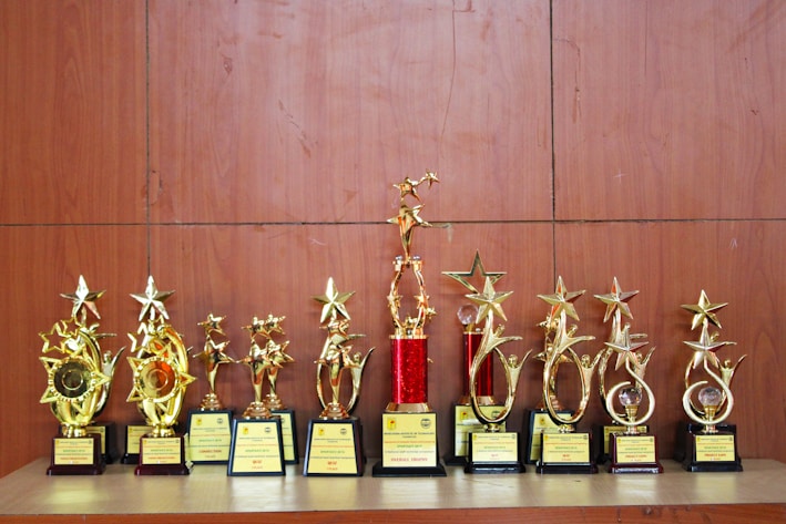 A collection of golden trophies with star designs displayed on a wooden surface. The trophies have various bases, some featuring red accents, and they appear to be awards given for special achievements.
