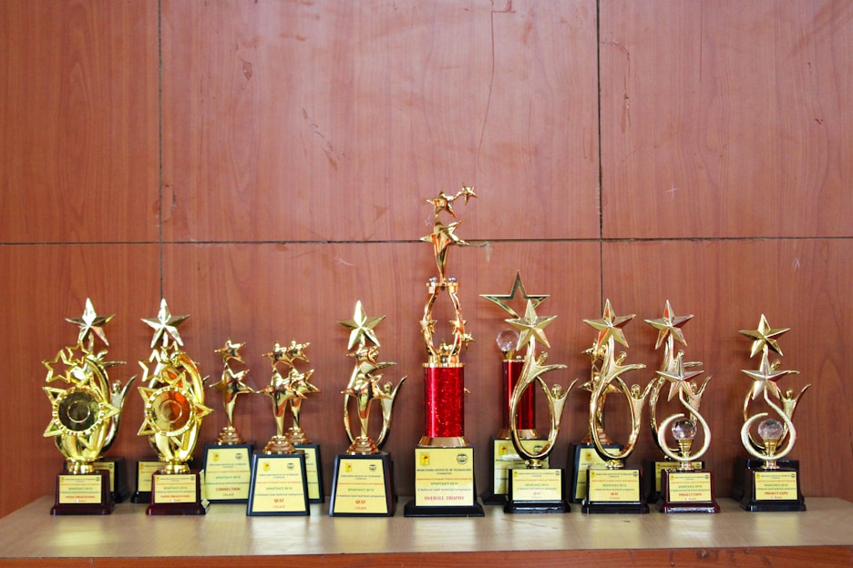 A collection of golden trophies with star designs displayed on a wooden surface. The trophies have various bases, some featuring red accents, and they appear to be awards given for special achievements.