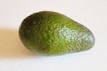 A single ripe avocado with a dark green, textured skin is placed on a light background. The avocado appears fresh and ready to eat, highlighting its rough surface and pear-like shape.