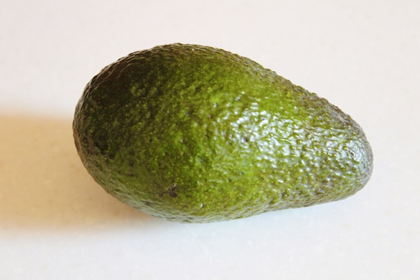 A single ripe avocado with a dark green, textured skin is placed on a light background. The avocado appears fresh and ready to eat, highlighting its rough surface and pear-like shape.
