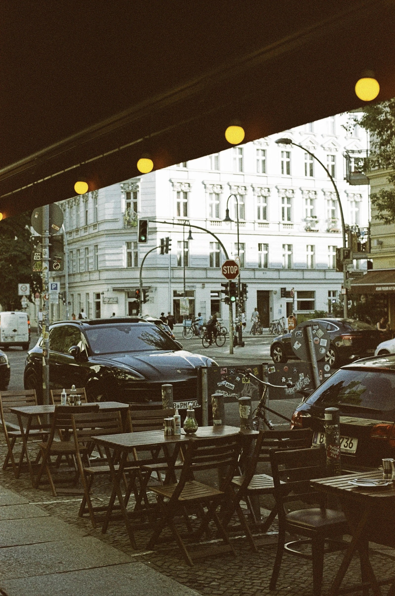 Travel photograph: Empty café tables sit under a dark awning on Schoenhauser Allee as cyclists and traffic pass beyond.