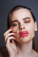 A woman with striking eye makeup and red lipstick smeared around her lips, touching her face with her hand. Her expression is serious and contemplative.