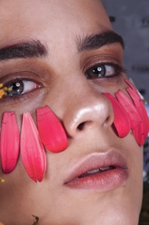 A close-up of a person's face with decorative red or pink flower petals placed on their cheeks. The person has bright, expressive eyes and neatly shaped eyebrows. The skin appears smooth and well-lit, highlighting the contrast between the petals and the skin.