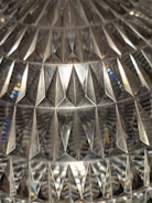 A detailed view of a textured surface with a geometric, diamond-like pattern, reflecting light and creating a variety of reflections and refractions. The surface appears to be glass or crystal, showcasing a complex array of triangular shapes and prismatic effects.