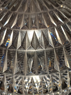 A detailed view of a textured surface with a geometric, diamond-like pattern, reflecting light and creating a variety of reflections and refractions. The surface appears to be glass or crystal, showcasing a complex array of triangular shapes and prismatic effects.