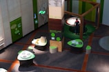 A modern exhibition space featuring interactive displays and informational panels predominantly in green tones. The area includes tables with digital screens and thematic displays about forestry and carbon footprint, and a person standing near a large, circular, interactive exhibit.