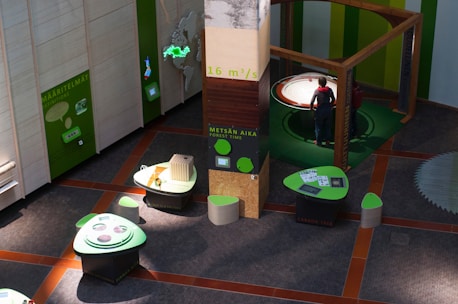 A modern exhibition space featuring interactive displays and informational panels predominantly in green tones. The area includes tables with digital screens and thematic displays about forestry and carbon footprint, and a person standing near a large, circular, interactive exhibit.