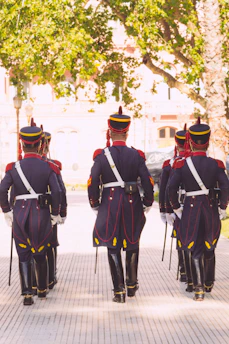 A group of soldiers dressed in formal military uniforms walks in formation along a pathway. They are wearing dark blue uniforms with red accents and tall hats, carrying swords. The setting appears to be outdoors in a park-like area, with green trees and sunlight filtering through the leaves.