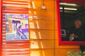 A vibrant painted wall features a colorful artwork of a couple dancing tango outside a café named 'Tortoni'. Next to it, a large window with a red frame reveals an elderly person indoors, reading a book while holding a cigarette.