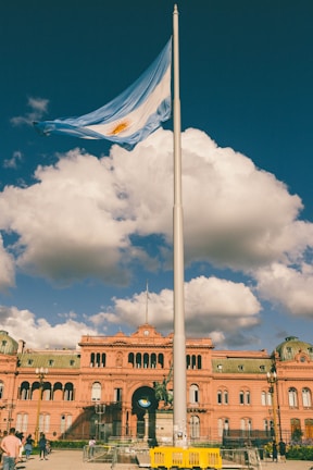 A large flagpole with the Argentine flag waving prominently in front of a historic pink building under a deep blue sky with fluffy clouds. Several people are scattered around the foreground. The architecture is ornate, with arches and detailed roof features.