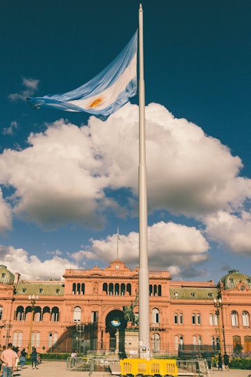 A large flagpole with the Argentine flag waving prominently in front of a historic pink building under a deep blue sky with fluffy clouds. Several people are scattered around the foreground. The architecture is ornate, with arches and detailed roof features.