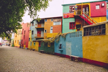 A vibrant street lined with colorful buildings painted in shades of red, yellow, blue, and green. The architecture includes bold geometric shapes and balconies. The cobblestone street adds a charming old-world feel, while the lush green tree on the left provides natural contrast to the vivid structures.