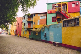 A vibrant street lined with colorful buildings painted in shades of red, yellow, blue, and green. The architecture includes bold geometric shapes and balconies. The cobblestone street adds a charming old-world feel, while the lush green tree on the left provides natural contrast to the vivid structures.