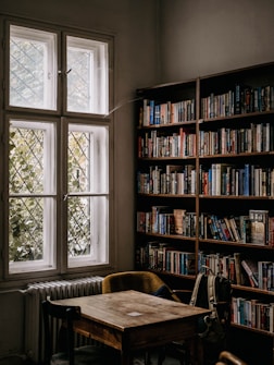 A cozy library corner featuring a wooden table and chairs placed near a window. The bookshelves are filled with various books, and a backpack is casually placed on a chair, suggesting a place for studying or quiet reading.