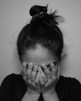 A person with hair tied in a bun covers their face with both hands, displaying an emotional moment. The lighting is subdued, emphasizing a sense of privacy and moodiness.