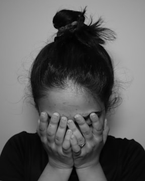 A person with hair tied in a bun covers their face with both hands, displaying an emotional moment. The lighting is subdued, emphasizing a sense of privacy and moodiness.