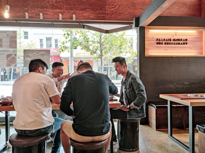Four people are seated around a table in a Korean BBQ restaurant, engaging in a lively conversation. The environment showcases a modern dining space with wooden interiors and large windows that allow in natural light. A sign with 'Palsaik Korean BBQ Restaurant' is visible on the wall.