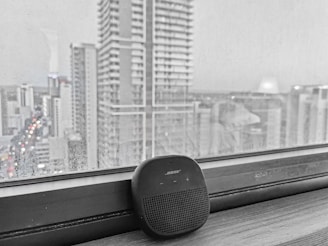 A sleek, modern smart speaker resting on a city apartment windowsill, with the skyline glowing softly in the background.