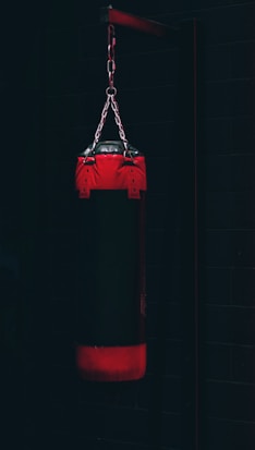 A boxing punching bag hangs from a metal chain against a dark background. The bag is predominantly black with red accents on the top and bottom. The environment appears dimly lit, creating a moody and intense atmosphere.