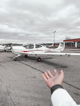 A small white aircraft with red accents is parked on a tarmac, with a partially extended hand in the foreground. The sky is overcast, and there are several other small planes and a building visible in the background.