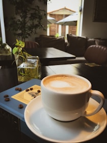 A white cup of coffee with a frothy top sits on a saucer atop a book with scattered coffee beans beside it. In the background, there are indoor plants and a cozy seating area featuring a couch. Natural light filters through large open windows, creating a warm, inviting atmosphere.