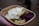 A cardboard container holds a serving of food, including cooked potatoes with herbs, slices of pickles, a dollop of creamy sauce, and a portion of red cabbage. The food is being held by a hand.