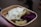 A cardboard container holds a serving of food, including cooked potatoes with herbs, slices of pickles, a dollop of creamy sauce, and a portion of red cabbage. The food is being held by a hand.