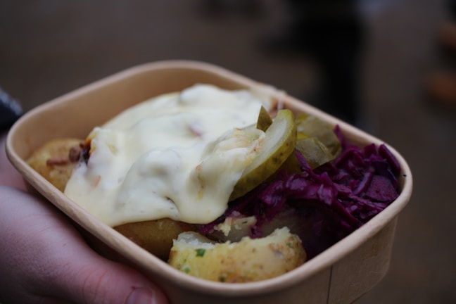 A cardboard container holds a serving of food, including cooked potatoes with herbs, slices of pickles, a dollop of creamy sauce, and a portion of red cabbage. The food is being held by a hand.