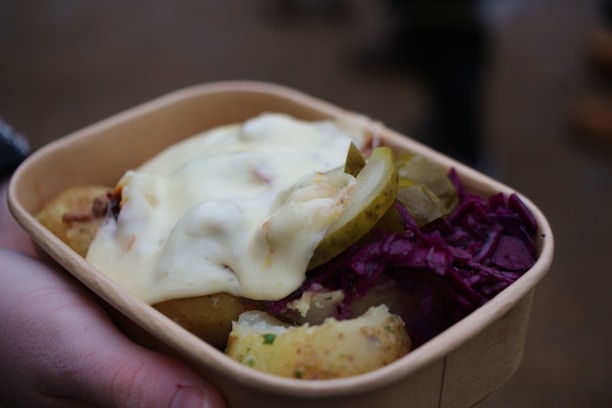 A cardboard container holds a serving of food, including cooked potatoes with herbs, slices of pickles, a dollop of creamy sauce, and a portion of red cabbage. The food is being held by a hand.