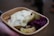 A cardboard container holds a serving of food, including cooked potatoes with herbs, slices of pickles, a dollop of creamy sauce, and a portion of red cabbage. The food is being held by a hand.