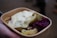 A cardboard container holds a serving of food, including cooked potatoes with herbs, slices of pickles, a dollop of creamy sauce, and a portion of red cabbage. The food is being held by a hand.