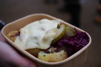 A cardboard container holds a serving of food, including cooked potatoes with herbs, slices of pickles, a dollop of creamy sauce, and a portion of red cabbage. The food is being held by a hand.