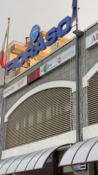 A building facade with a large sign reading 'FIORASO' in bold blue letters. Below the sign are various company logos and advertisements. There are flags on poles, and a digital clock display showing the temperature. The architectural style includes a curved awning and horizontal slats over large windows.