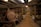 A dimly lit warehouse filled with rows of stacked wooden barrels, likely containing whiskey. The barrels are organized neatly on shelves, and the faded lettering on some indicates their age and origin. The space has a rustic, industrial feel with metal beams and concrete floors.