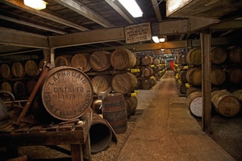 A dimly lit warehouse filled with rows of stacked wooden barrels, likely containing whiskey. The barrels are organized neatly on shelves, and the faded lettering on some indicates their age and origin. The space has a rustic, industrial feel with metal beams and concrete floors.