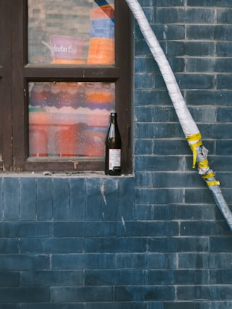 A weathered dark blue brick wall with a window. Inside the window is a collection of stacked colorful plastic containers, and outside the window on the ledge stands a dark glass bottle with a label in a foreign language. A large, white insulated pipe runs vertically alongside the window and is secured with yellow tape.