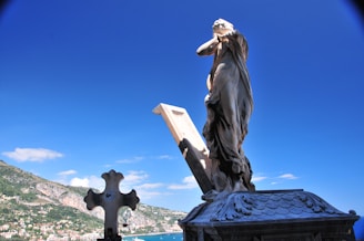 A stone statue of a robed figure stands on a rooftop with an open book beside it. The background features a clear blue sky and a lush, mountainous landscape overlooking a coastal town.
