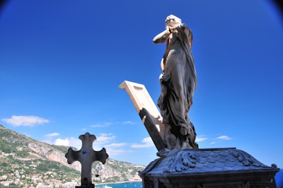 A stone statue of a robed figure stands on a rooftop with an open book beside it. The background features a clear blue sky and a lush, mountainous landscape overlooking a coastal town.