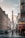 A bustling Parisian street scene with the Eiffel Tower visible in the distance. People are walking and cycling along the street lined with classic European buildings. A café with a red canopy and pedestrians crossing the road add to the lively atmosphere.